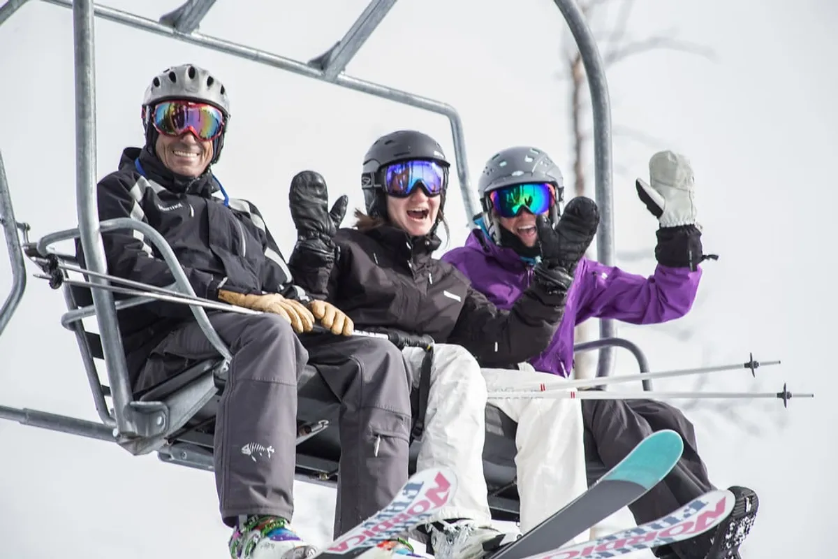 Three skiers ride a chairlift together, surrounded by a snowy landscape. The scene is cheerful and social, highlighting the communal aspects of ski trips—shared laughter, anticipation, and the thrill of a day on the slopes.
