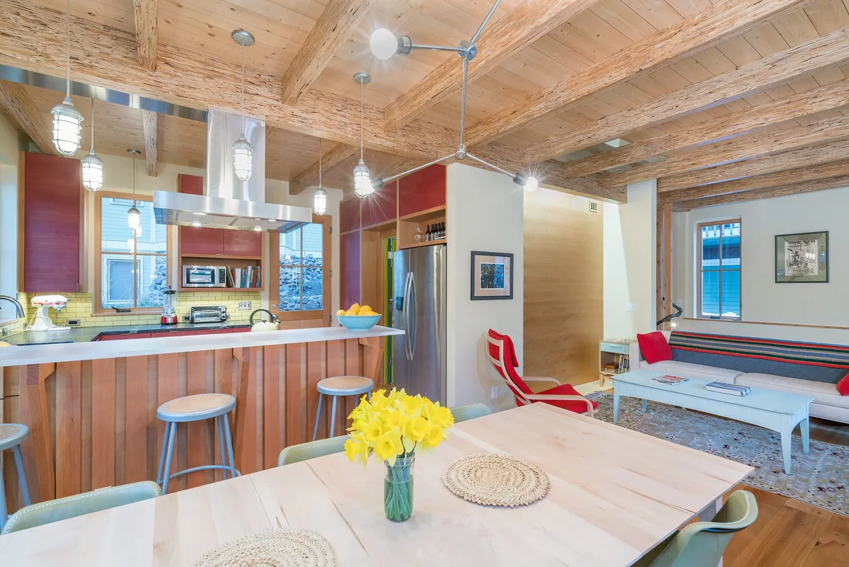 This interior image showcases an open-plan communal area that seamlessly connects the kitchen island, a dining table adorned with flowers, and a cozy living area with comfortable seating. Large windows flood the entire space with natural sunlight, underscoring the sense of warmth and connection described in the section.
