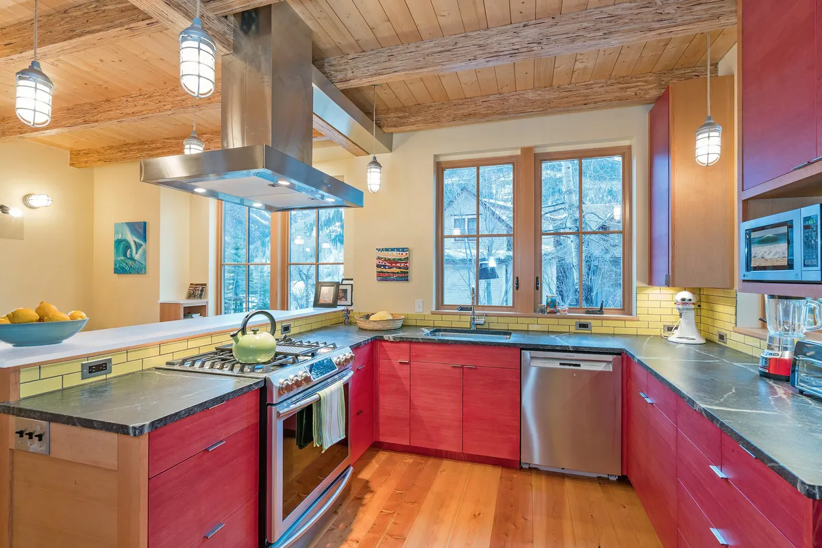 The photo focuses on a stylish kitchen featuring a central island with a sink, stainless steel appliances, striking red cabinetry, and large windows. The space is clearly designed for socializing and preparing meals, with sunlight streaming in and modern, eco-conscious design elements evident.
