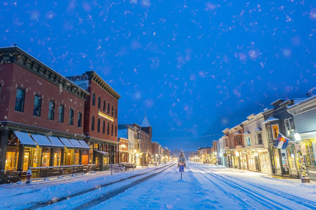 A snow-covered main street lined with historic buildings, warmly lit by streetlights. A large Christmas tree glows at the center, and people are seen strolling and enjoying the festive ambiance. The scene evokes Telluride’s lively and walkable downtown atmosphere, especially during the winter season.
