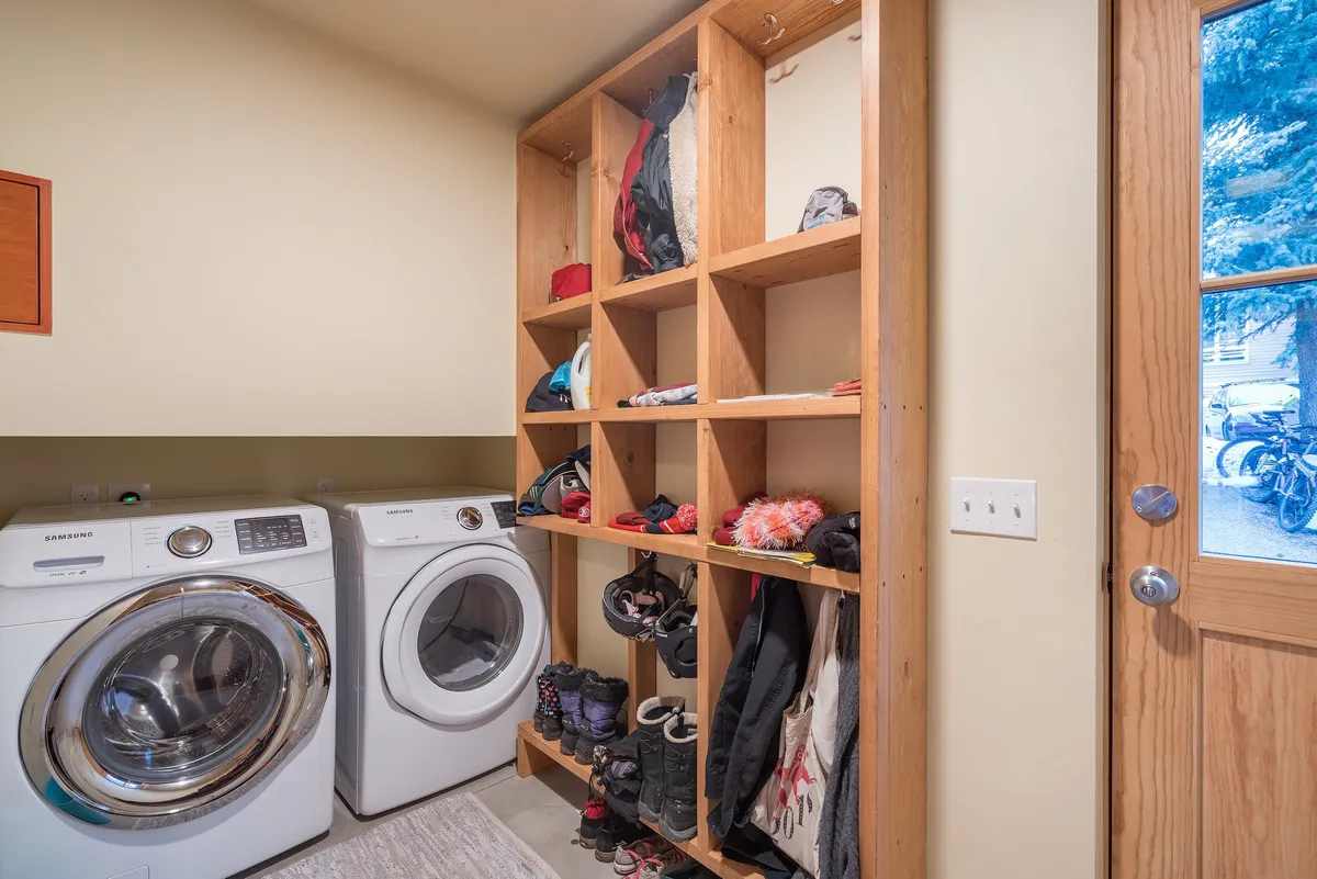 A practical and tidy laundry area featuring a modern washing machine, organized shelving stocked with supplies, and a welcoming front door. The space is clearly designed for family use, making chores convenient after outdoor adventures.