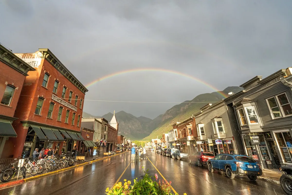 A cheerful street scene in Telluride's historic downtown, featuring colorful buildings, a rainbow arching overhead, wet streets from a recent rain, people strolling, and bicycles parked along the sidewalk. The setting captures the vibrancy and walkability of the area, as well as the family-friendly, small-town charm.