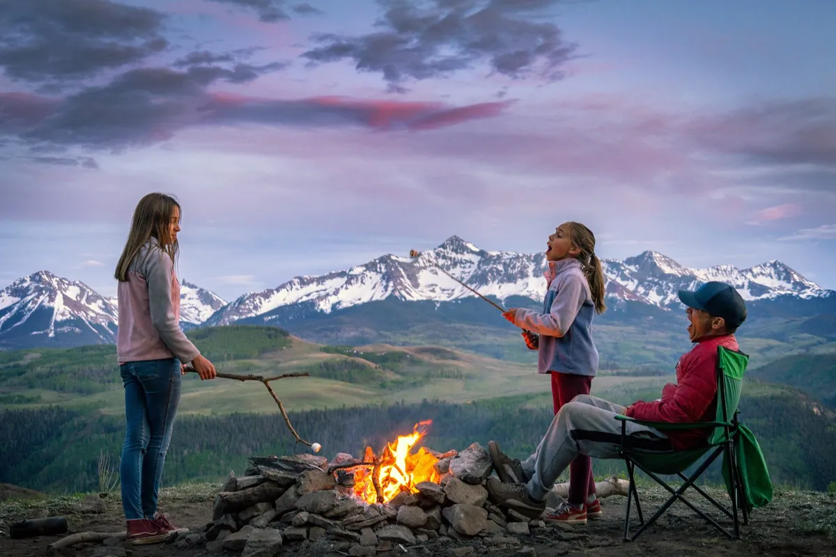 A nostalgic, joyful campfire setting with children roasting marshmallows against a mountain backdrop. The atmosphere is warm and inviting, with a group gathered to enjoy a classic family experience at sunset.