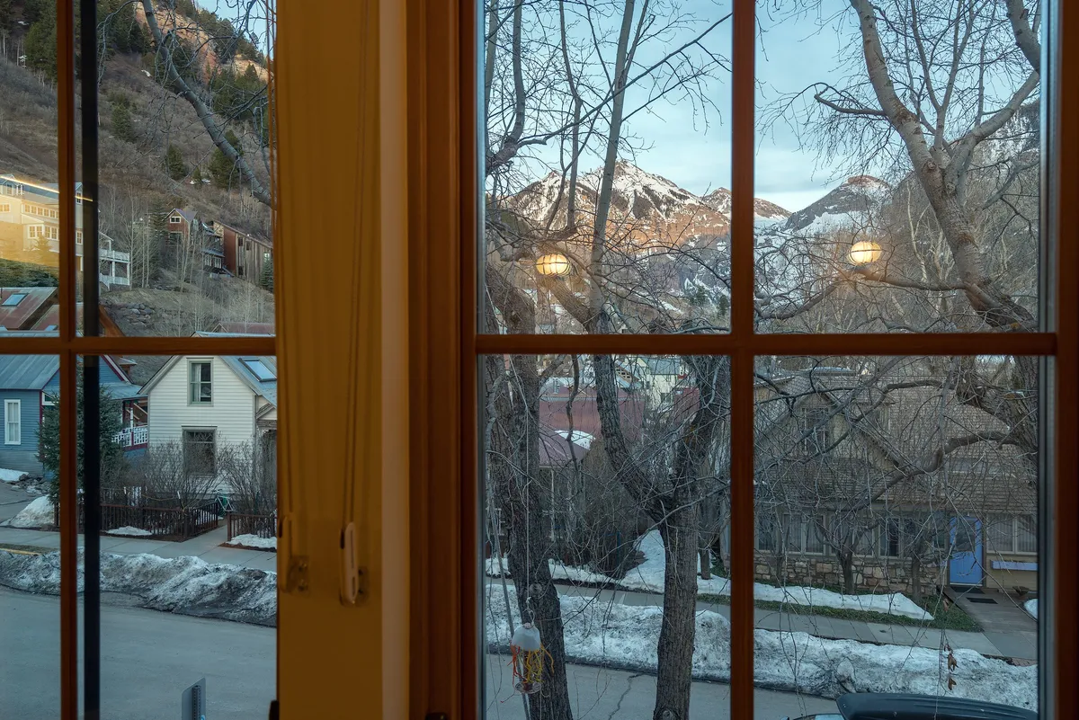 A tranquil view from indoors looking out toward snow-capped mountains and trees, with nearby houses visible. The peaceful setting suggests a comfortable retreat where guests can relax and enjoy the scenery after a night out in Telluride.