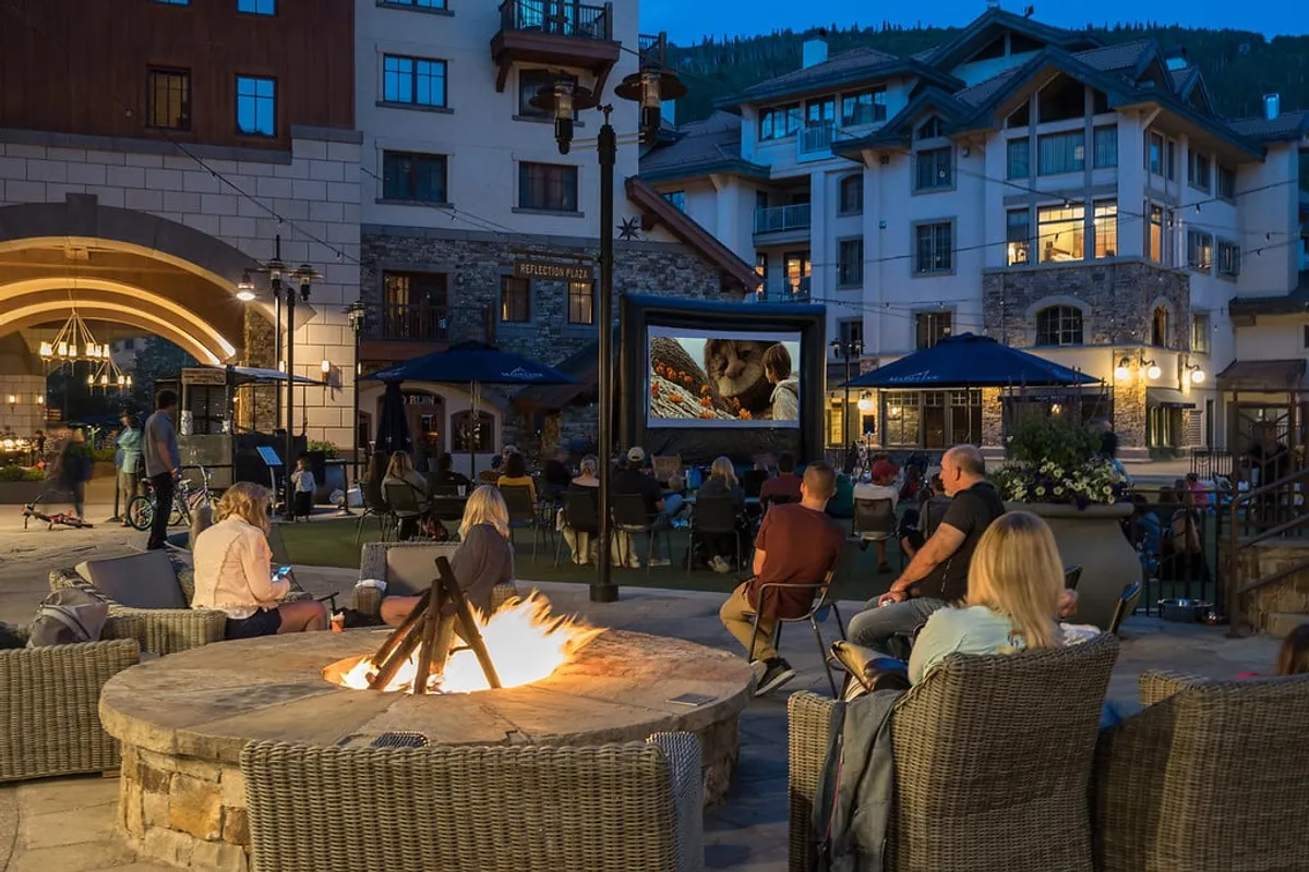 An inviting outdoor scene featuring a fire pit surrounded by comfortable seating, an outdoor movie screen, and architectural details of the building. The area is aglow with warm lighting, suggesting a perfect setting for gathering after an evening out.