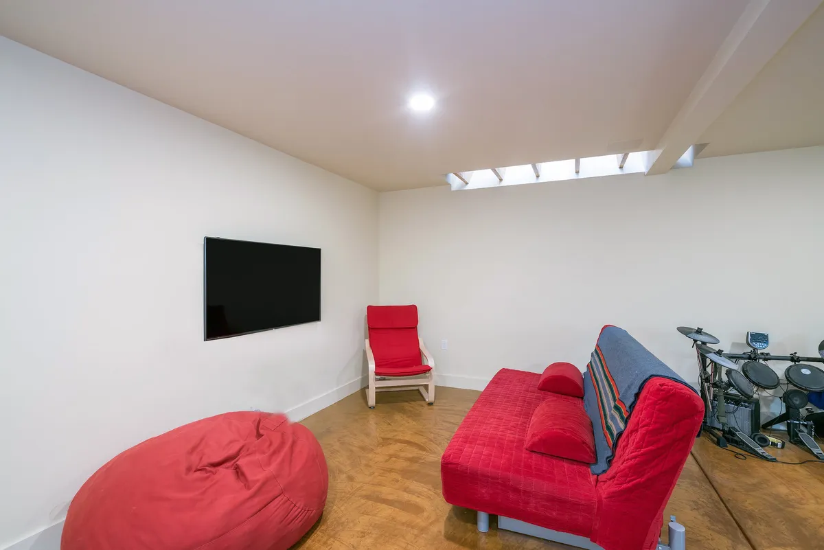 This image features a vibrant basement lounge with a red couch, bean bag chair, TV, red chair, and musical instruments. The space is inviting and playful, designed for activities such as watching movies, playing video games, relaxing, and music practice. The arrangement suggests a flexible layout with comfortable seating options.