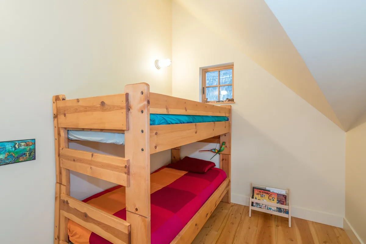 A playful bunk room complete with a bunk bed adorned in colorful bedding, a small bookshelf, and a window that lets in gentle morning light. The space is clearly designed for children, inviting both sleep and play.