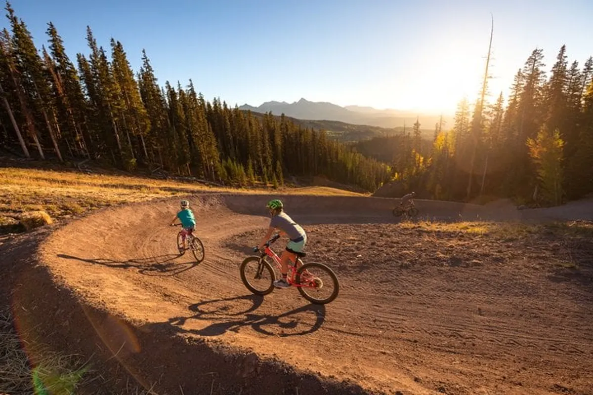 This exterior shot captures mountain bike riders navigating a curved dirt trail against the backdrop of a golden sunset. The surrounding scenery is lush and vibrant, evoking a sense of adventure and connection to nature. The warm light and visible camaraderie among the riders embody outdoor activity and shared experiences.