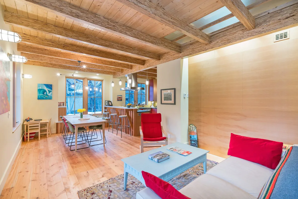 A welcoming interior scene with exposed wooden beams, red accents, a dining table decorated with flowers, and a plush sofa set within a cozy living space. The arrangement invites conversation and relaxation, with a sense of warmth and artistic flair.