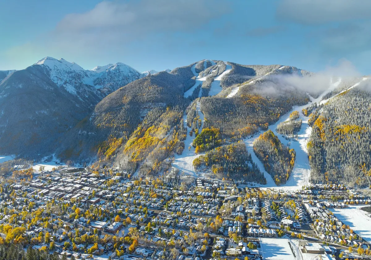 A breathtaking exterior view of Telluride’s snow-covered mountains and ski slopes, with colorful autumn trees framing the town below. The scene encapsulates both winter and autumn activities, from skiing to hiking, and the beauty of the changing seasons.