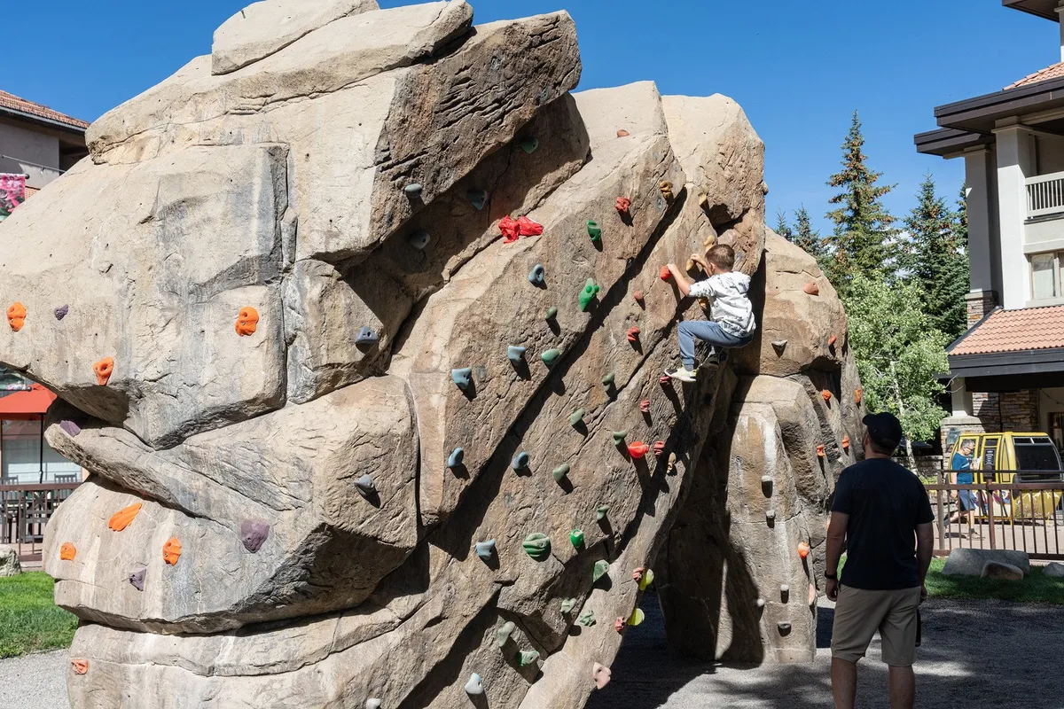 An outdoor climbing wall scene with a child actively climbing while an adult watches and offers encouragement below. The backdrop includes natural elements, reinforcing a focus on outdoor play within a mountain environment.