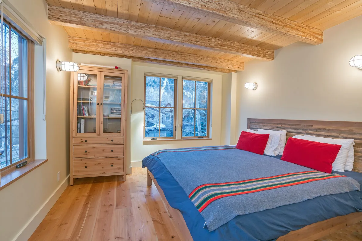 A serene, sunlit bedroom with a comfortable bed accented by colorful throw pillows, a sturdy wooden cabinet, and large windows that fill the space with natural light. The environment exudes relaxation and hominess suitable for families.