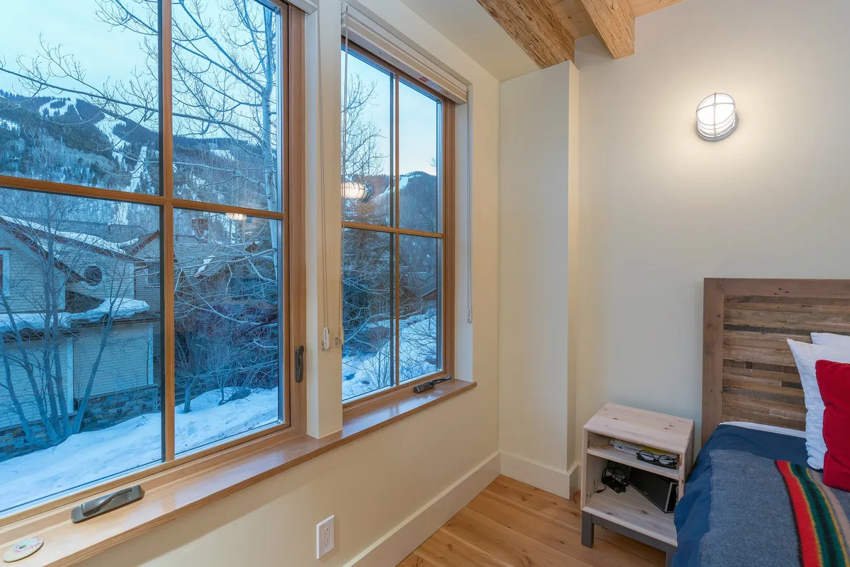 A tranquil bedroom with a wooden bed frame positioned near large windows, complemented by a simple nightstand. Through the windows, snow blankets the landscape, suggesting a serene winter scene and offering beautiful mountain views to wake up to.