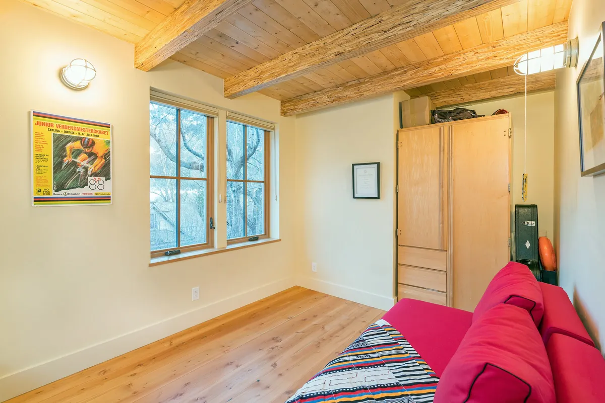 A modern, cozy living space featuring a red couch, large windows letting in natural light, and exposed wooden beams. The room feels spacious and adaptable, suitable for gatherings or quiet moments alike.