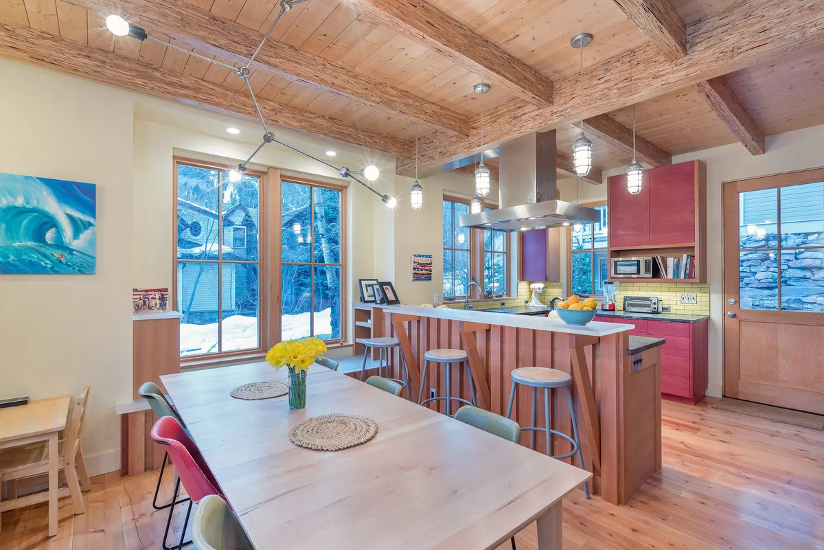 A sunlit, open-concept kitchen and dining area featuring a wooden dining table with fresh flowers, a sleek kitchen island, exposed wooden beams, and large windows that flood the space with natural light and offer tranquil views.